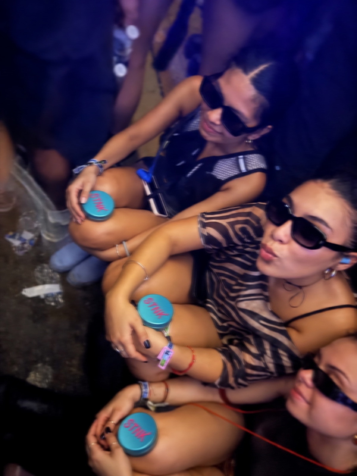 Friends sitting on the floor at a festival, each with a teal STNK tin on their knee