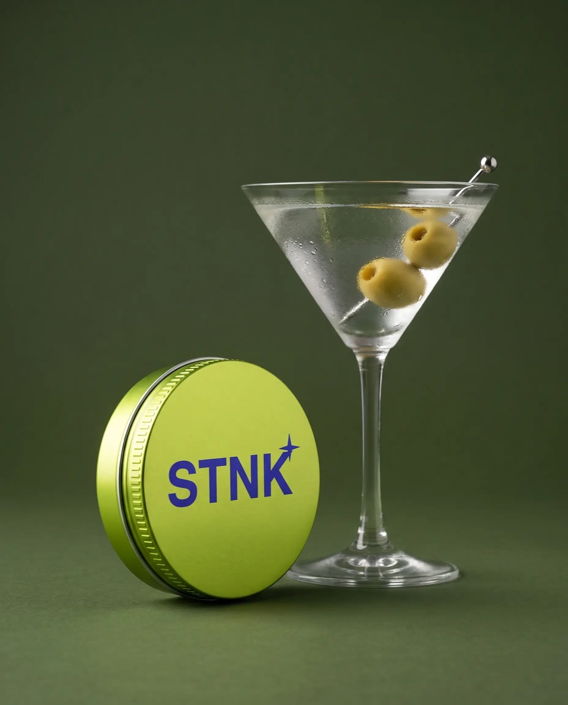 The lime Dirty Martini STNK tin beside a martini with olives